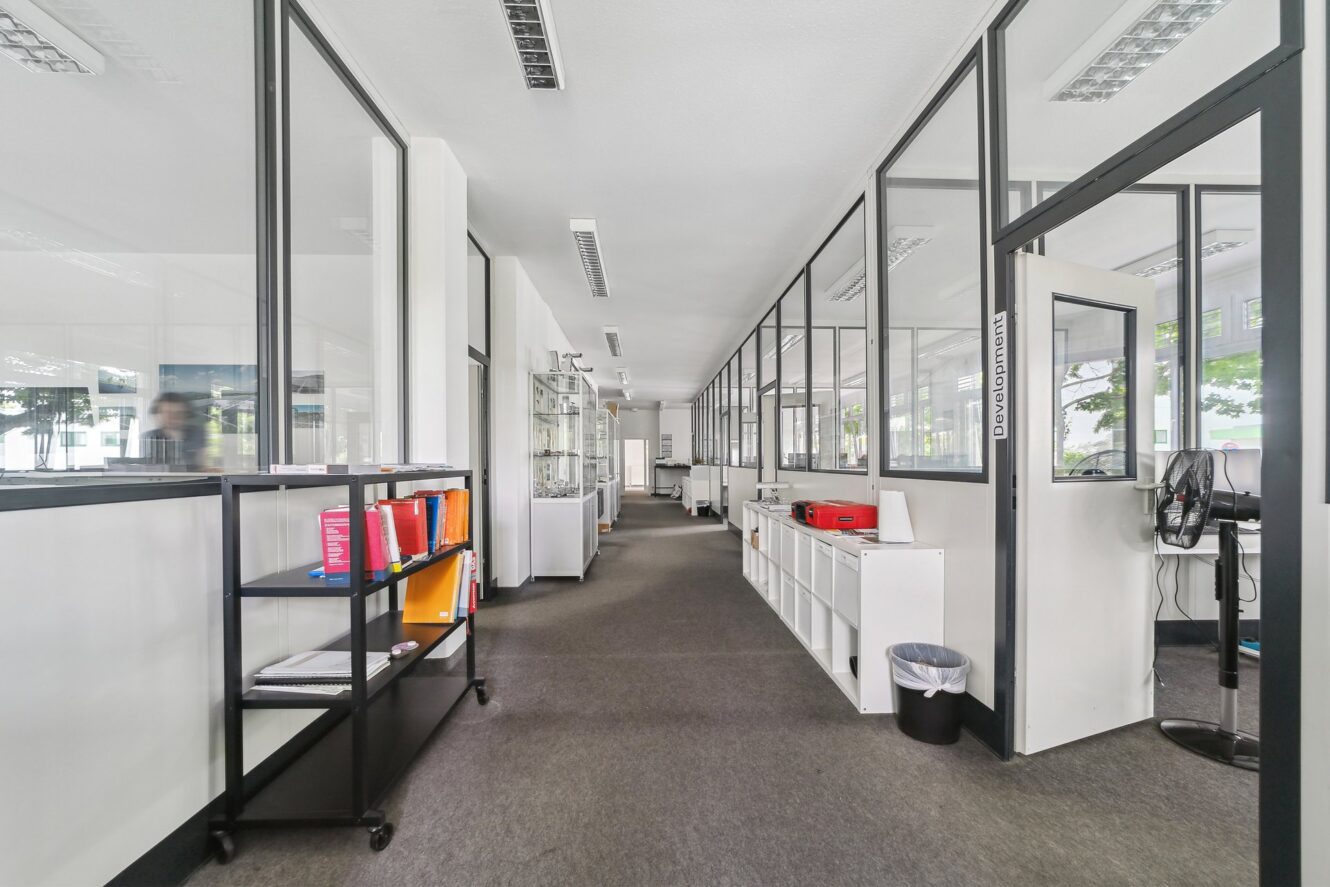 Moderner Büroflur mit Glaswänden, Regalen mit Büchern und Büromaterial sowie einem Schreibtisch mit Computer. Der Raum ist gut beleuchtet und geordnet.