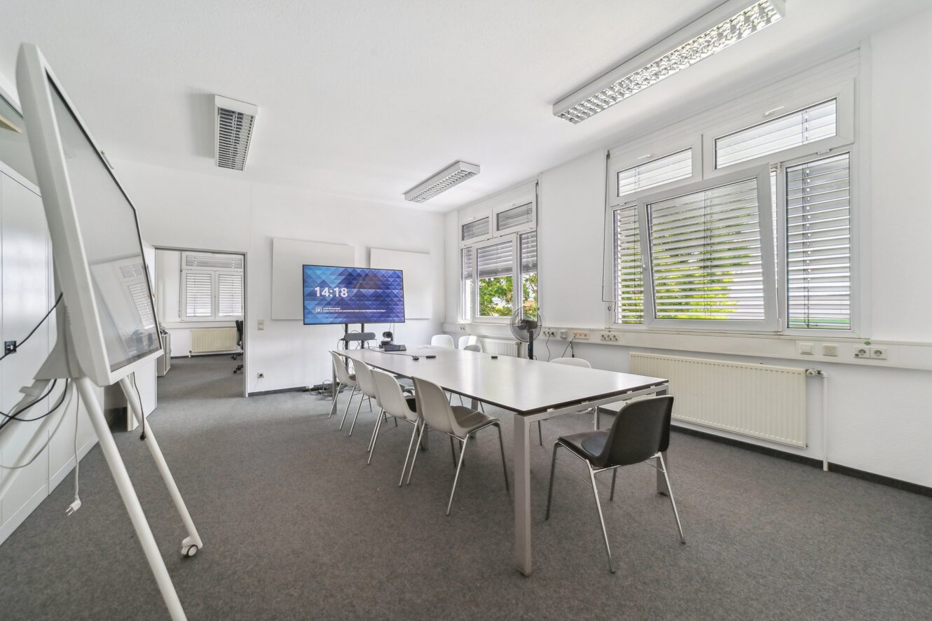 Moderner Konferenzraum mit einem langen Tisch, weißen Stühlen, einem großen Bildschirm, der die Uhrzeit anzeigt, einem Whiteboard und großen Fenstern mit Jalousien.
