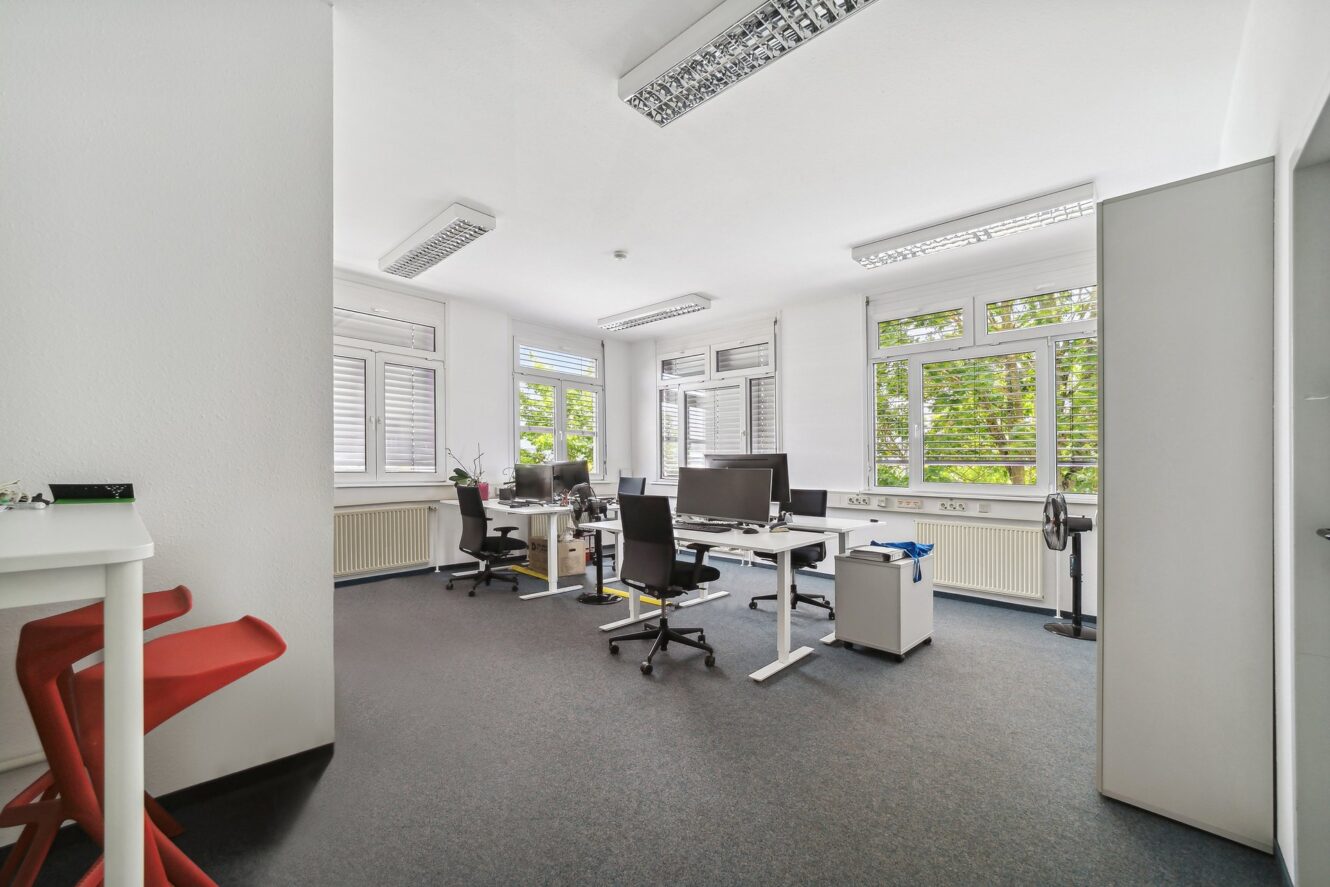 Heller Büroraum mit mehreren Schreibtischen, Computern, Stühlen und großen Fenstern, die viel Tageslicht hereinlassen; links stehen zwei rote Hocker an einem weißen Tresen.