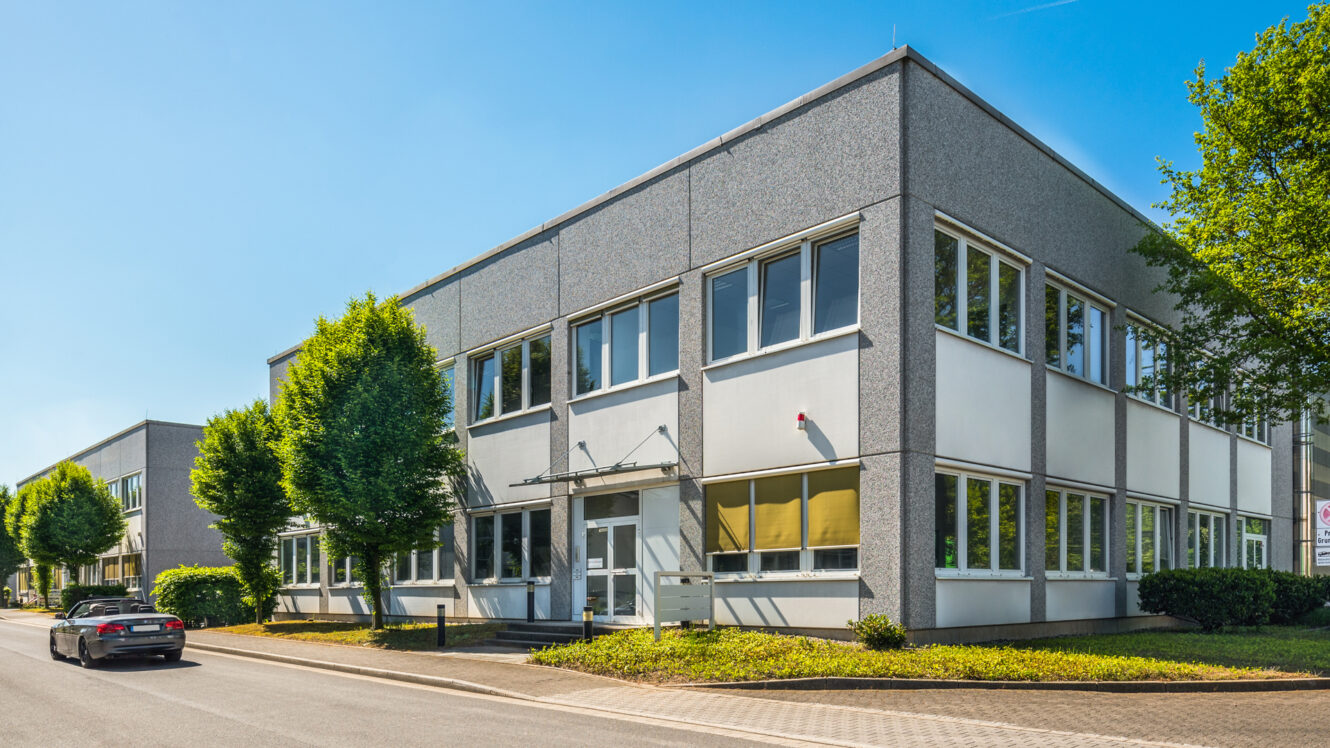 Ein modernes, zweistöckiges Bürogebäude mit großen Fenstern an einem sonnigen Tag, direkt an einer Straße mit einem geparkten Auto und Bäumen.