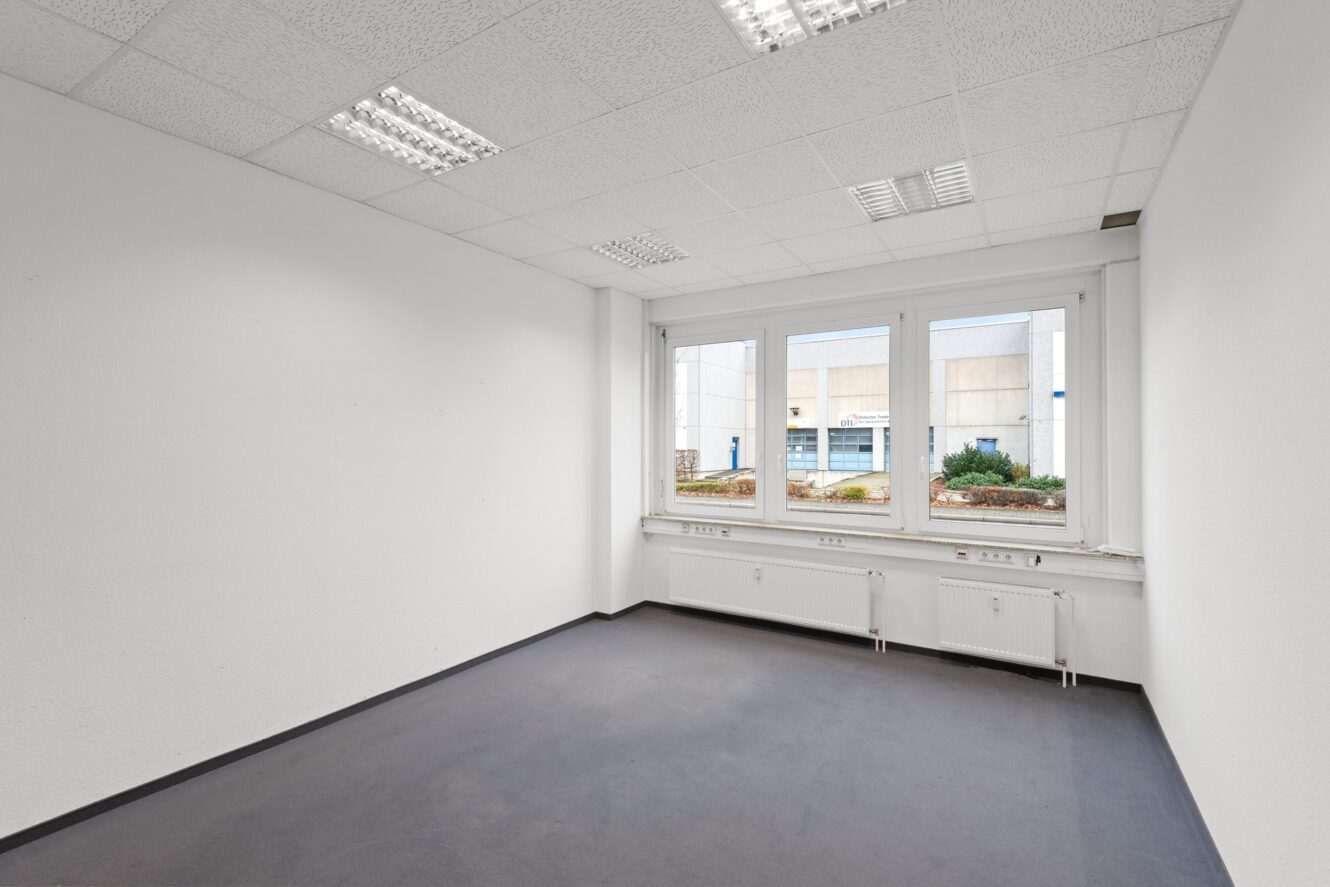 Leerer Büroraum mit weißen Wänden, grauem Boden, großen Fenstern und fluoreszierenden Deckenleuchten. Die Fenster geben den Blick auf ein Gebäude im Außenbereich und einige Grünanlagen frei.