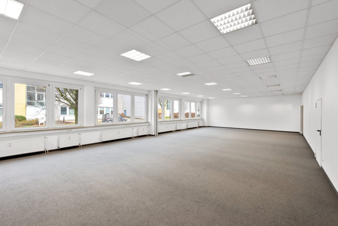 Ein großer, leerer Büroraum mit grauem Teppich, weißen Wänden, einer Fensterreihe auf einer Seite und an der Decke angebrachten Leuchtstoffröhren.