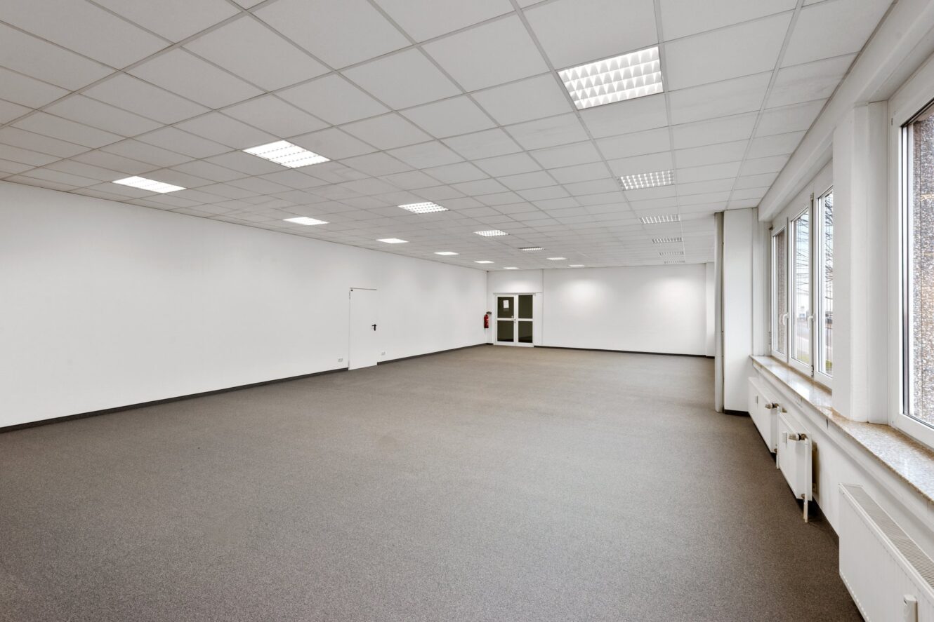 Ein leerer Büroraum mit weißen Wänden, einer Gitterdecke mit Leuchtstoffröhren, grauem Teppichboden und großen Fenstern an einer Wand.