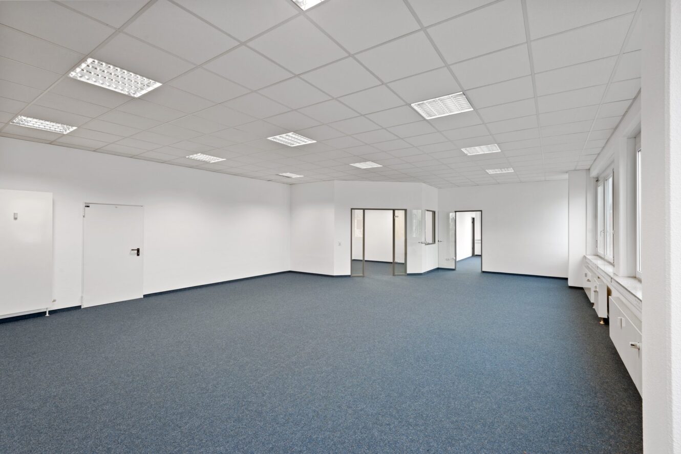 Leerer Büroraum mit blauem Teppich, weißen Wänden, Deckenleuchten, Fenstern auf der rechten Seite und einigen Innentüren und Glastrennwänden auf der Rückseite.