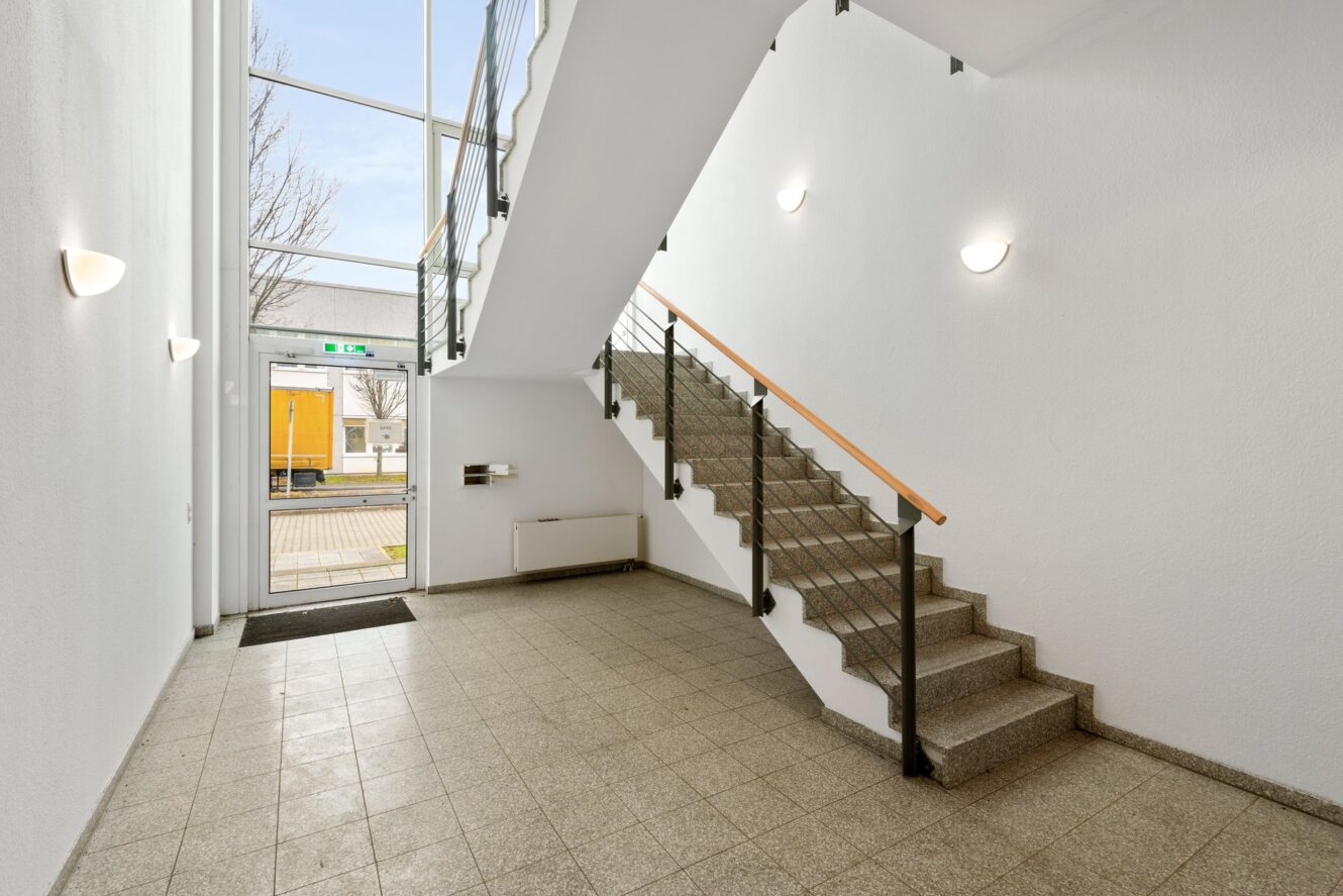 Ein sauberes, gut beleuchtetes Treppenhaus mit gefliestem Boden, weißen Wänden und Metallgeländern. Große Fenster sorgen für natürliches Licht und einen Blick auf die Straße und das Gebäude draußen.