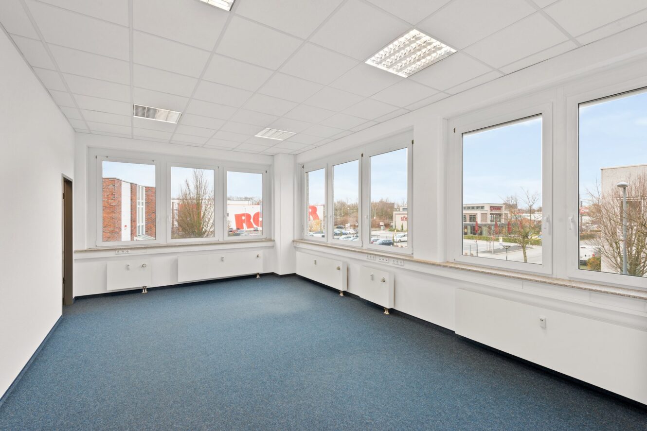 Leerer Büroraum mit blauem Teppich, weißen Wänden, großen Fenstern und Leuchtstoffröhrenbeleuchtung. Draußen sind städtische Gebäude und geparkte Autos zu sehen.