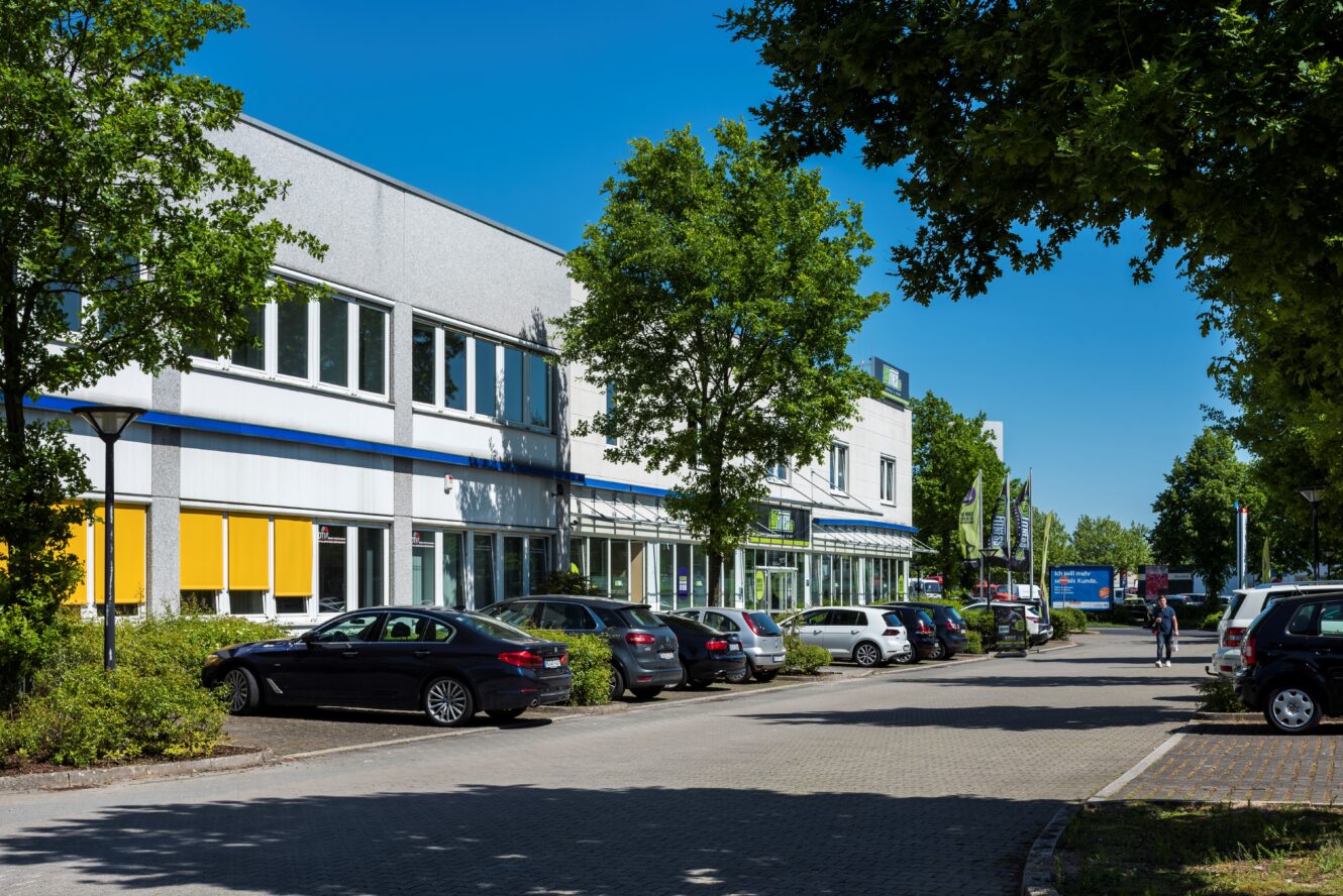 Eine Reihe geparkter Autos säumt eine Straße neben einem modernen Bürogebäude mit Bäumen und blauem Himmel über dem Kopf.