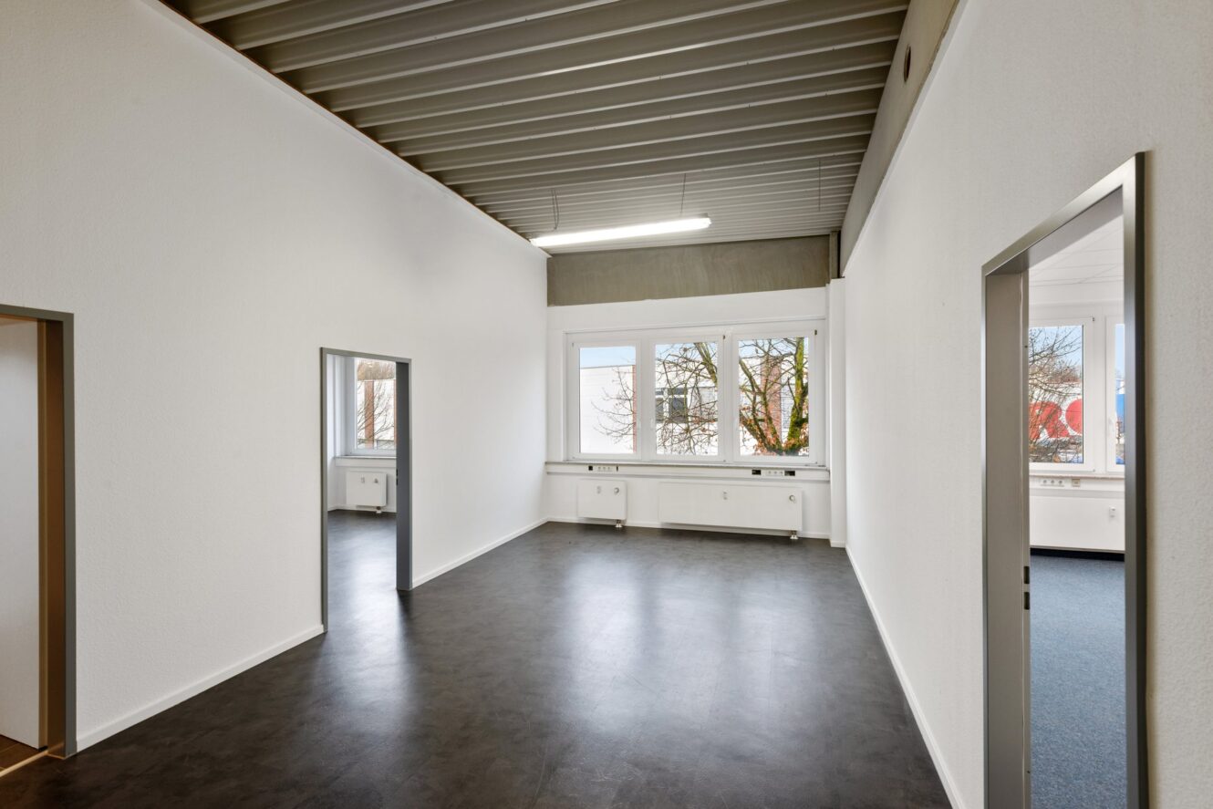 Leerer, moderner Büroraum mit weißen Wänden, dunklem Fußboden, großen Fenstern und zwei offenen Türen, die zu angrenzenden Räumen führen.