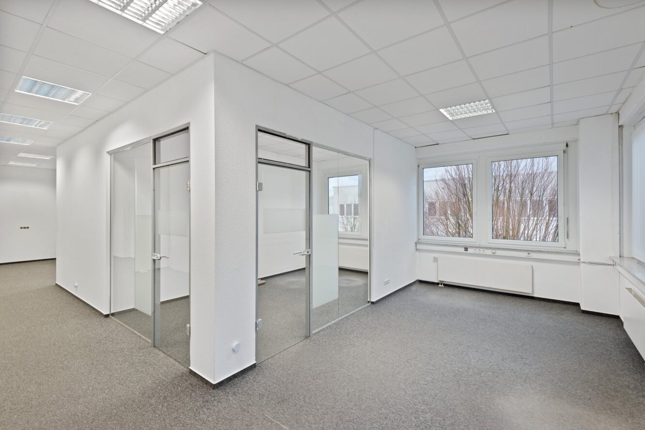 Moderne, leere Büroräume mit Glaswänden, weißen Wänden, grauem Teppich und großen Fenstern, die viel Tageslicht hereinlassen.
