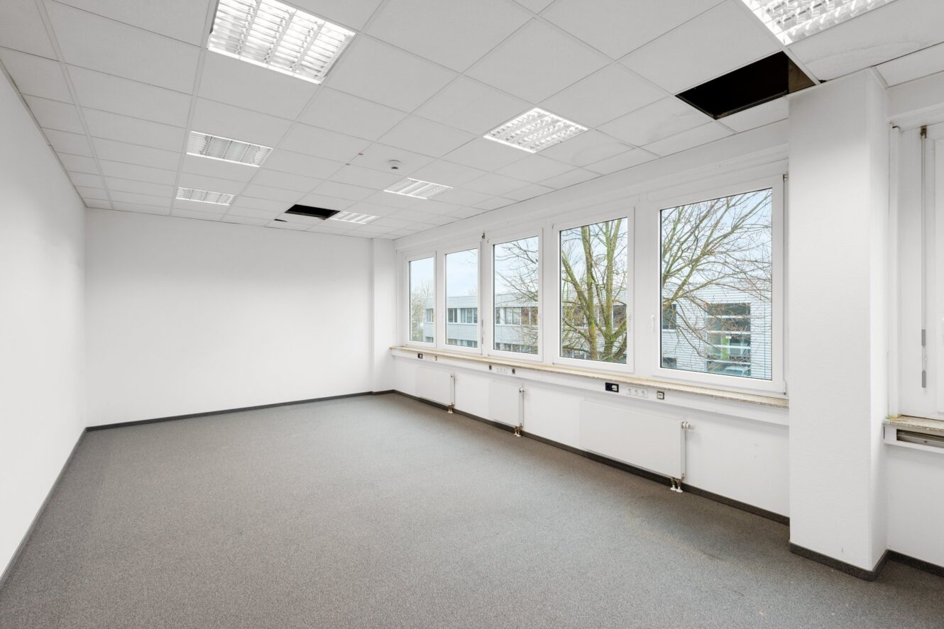 Leerer Büroraum mit weißen Wänden, Teppichboden, großen Fenstern, Leuchtstoffröhren an der Decke und einigen fehlenden Deckenplatten.