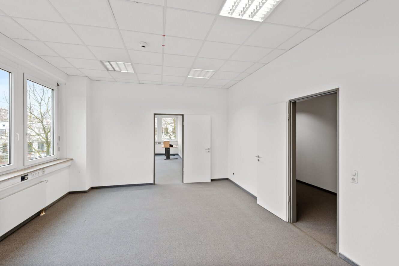 Leerer Büroraum mit weißen Wänden, grauem Teppich, Deckenleuchten, großem Fenster auf der linken Seite und offenen Türen, die zu angrenzenden Räumen führen.