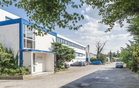 Ein weißes Industriegebäude mit blauer Verkleidung, umgeben von Bäumen und geparkten Autos, unter einem teilweise bewölkten Himmel.