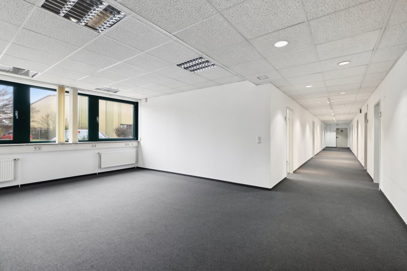 Ein großer, leerer Büroraum mit weißen Wänden, grauem Teppich, Deckenleuchten, Fenstern auf der linken Seite und einem Flur mit mehreren Türen auf der rechten Seite.