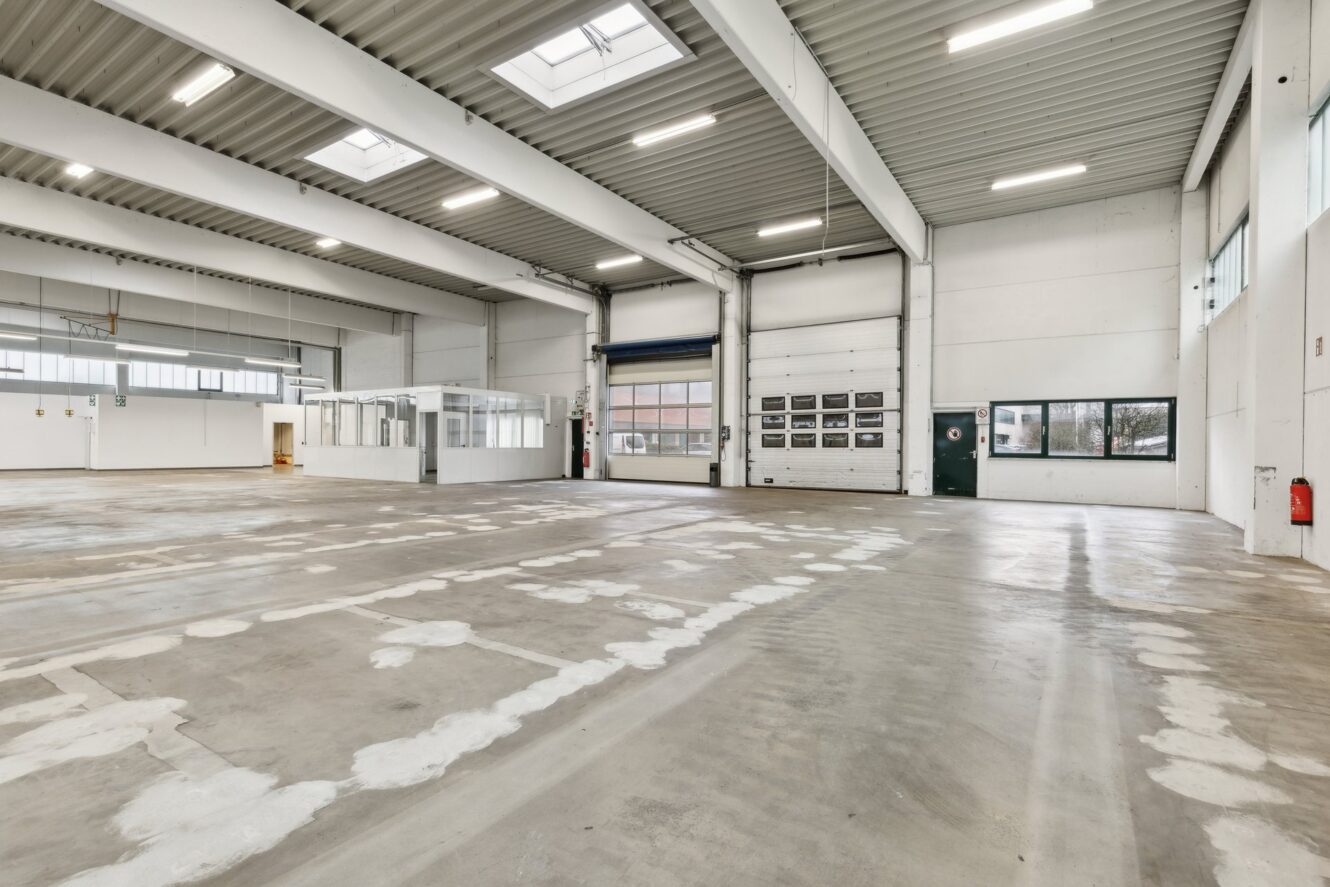 Geräumiges, leeres Industrielager mit großen Fenstern, Oberlichtern, Betonboden, hohen Decken und einem kleinen Bürobereich auf der linken Seite.