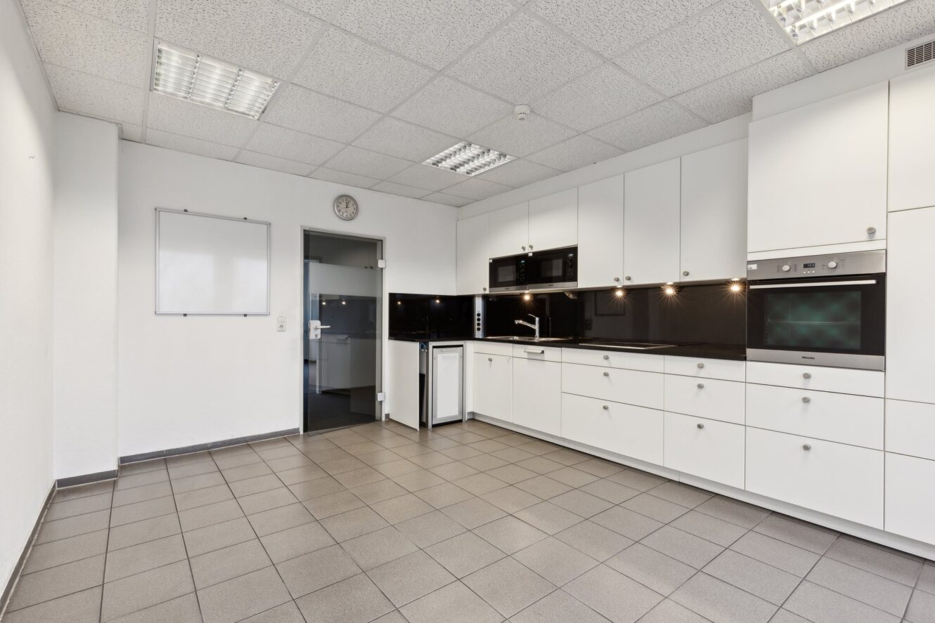 Moderne Küche mit weißen Schränken, schwarzen Arbeitsplatten, Einbaugeräten und grau gefliestem Boden. Ein Whiteboard und eine Uhr sind an der Wand neben einer Tür angebracht.