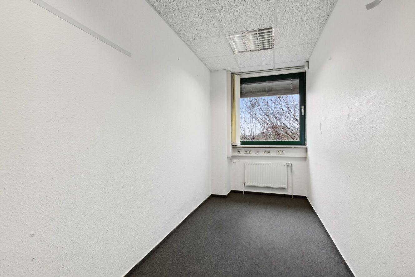 Kleiner, leerer Büroraum mit weißen Wänden, dunklem Teppich, einem Fenster mit einem Heizkörper darunter und einer Leuchtstoffröhre an der Decke.