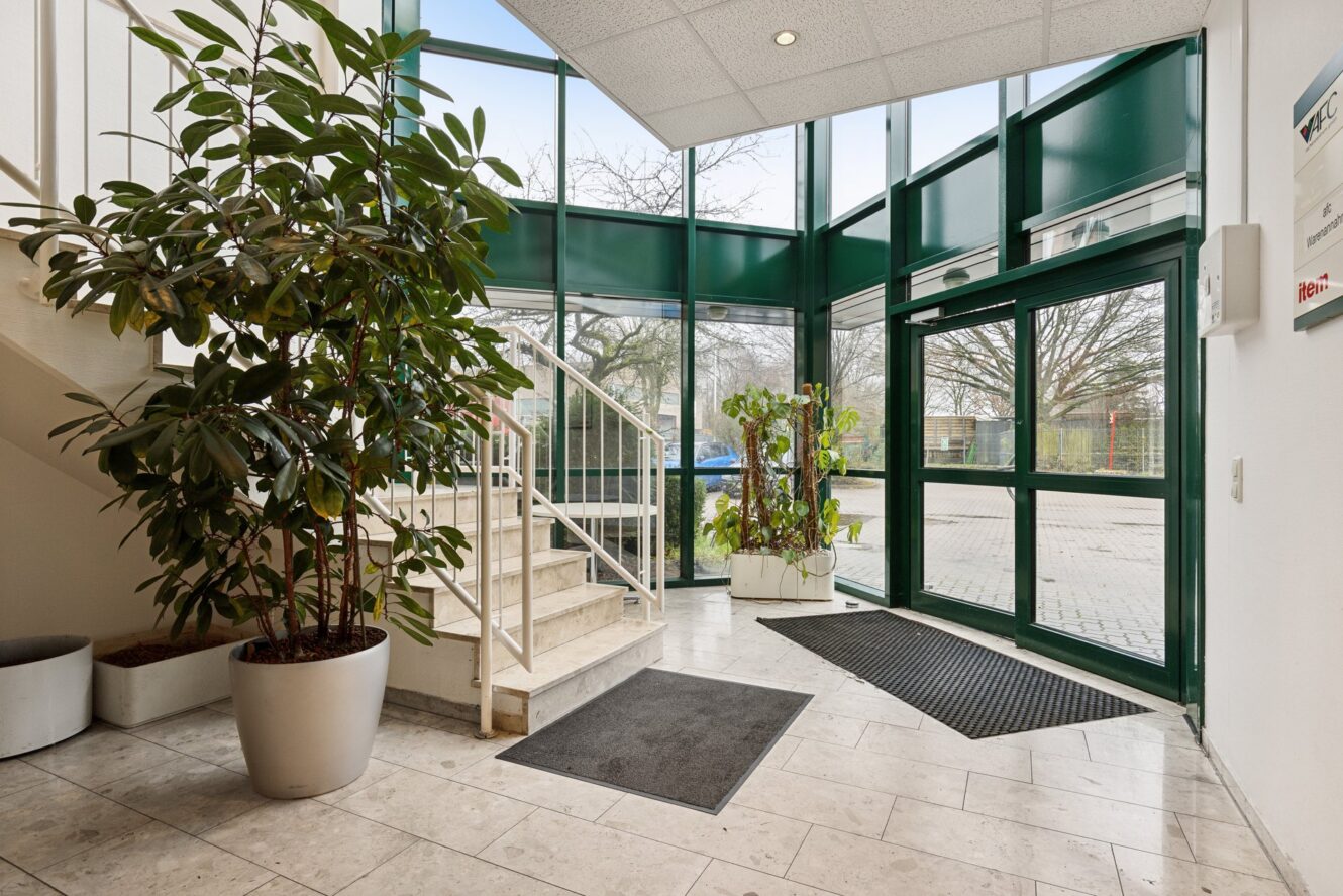 Helle Bürolobby mit großen Fenstern, Topfpflanzen, Fliesenboden, Treppe auf der linken Seite und doppelten Glastüren, die nach draußen führen.