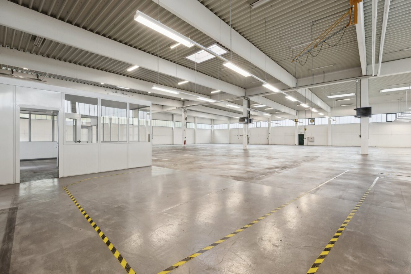 Geräumiges, leeres Industrielager mit hohen Decken, weißen Wänden, großen Fenstern und Deckenbeleuchtung; gelbe Warnstreifen markieren Bereiche auf dem Betonboden.