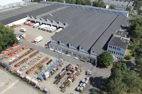 Luftaufnahme eines Industrielagers mit einem großen grauen Dach, angrenzendem Parkplatz, Lieferwagen und organisierter Außenlagerung von Paletten und Baumaterialien.