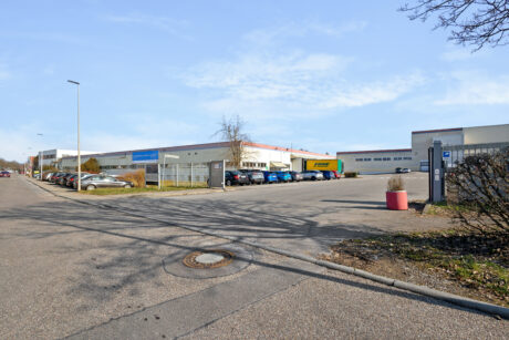 Breite Straßenansicht eines Industriegebiets mit Lagerhallen, geparkten Autos, einem DHL-LKW und klarem Himmel im Hintergrund.