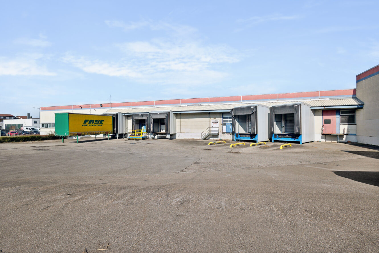 Ein gelber Frachtanhänger ist an einem klaren Tag an einer Laderampe vor einem großen Industrielager geparkt.