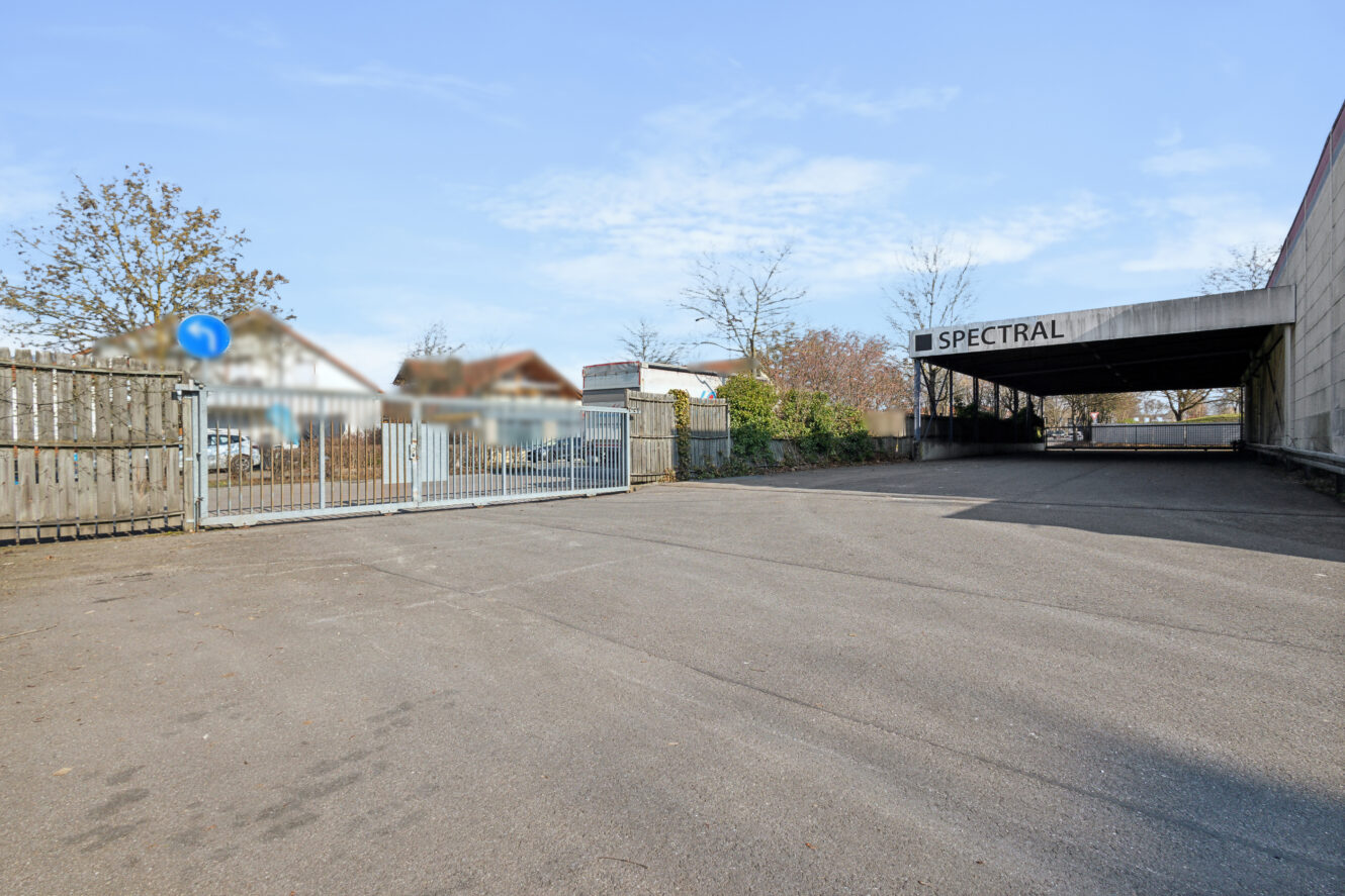 Gepflasterte Zufahrt, die zu einem bewachten Eingang und einem Gebäude mit SPECTRAL-Schildern führt, umgeben von Bäumen und Häusern im Hintergrund.