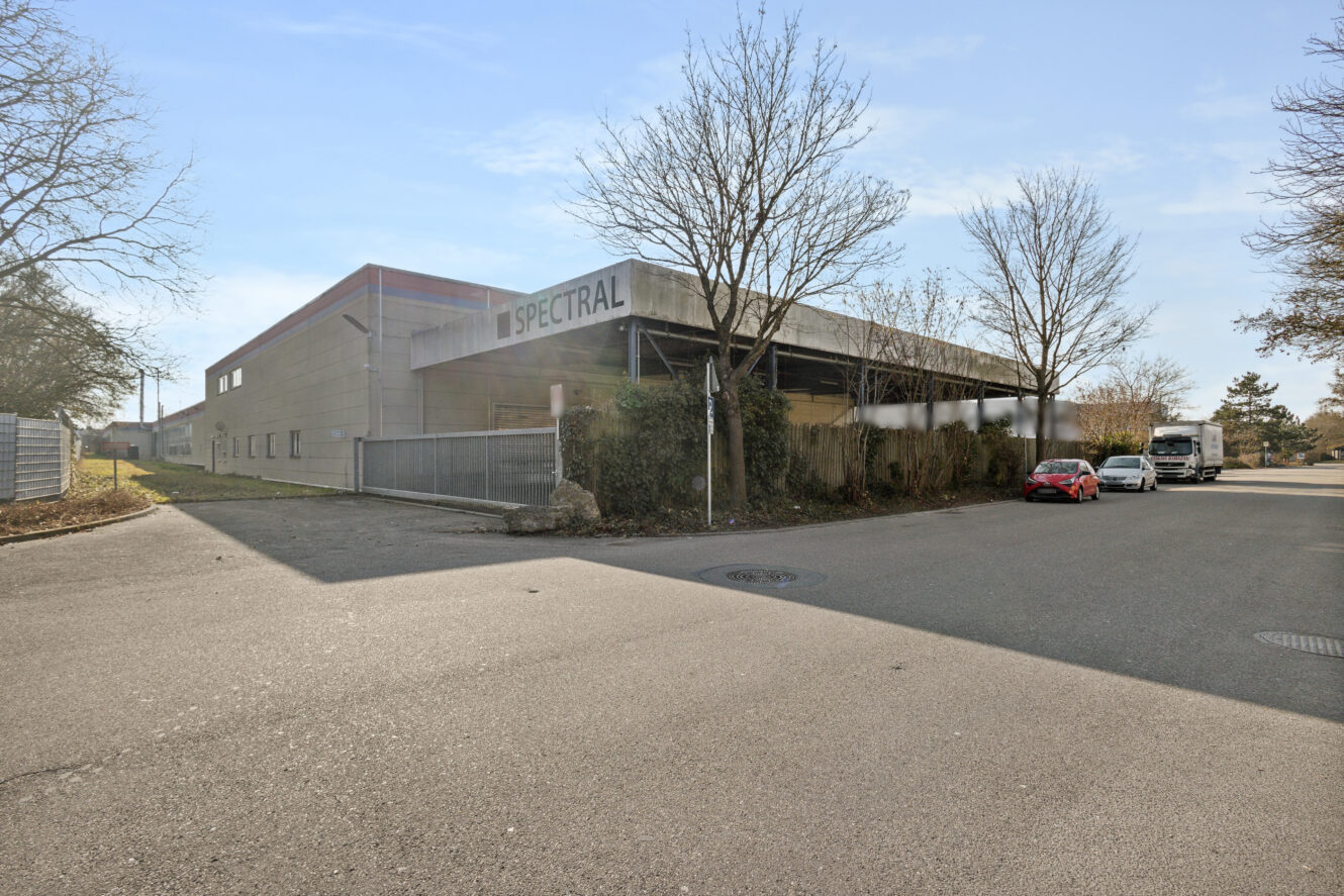 Ein großes Industriegebäude mit dem Zeichen SPECTRAL, umgeben von kahlen Bäumen, einem Zaun und ein paar geparkten Fahrzeugen an einem sonnigen Tag.