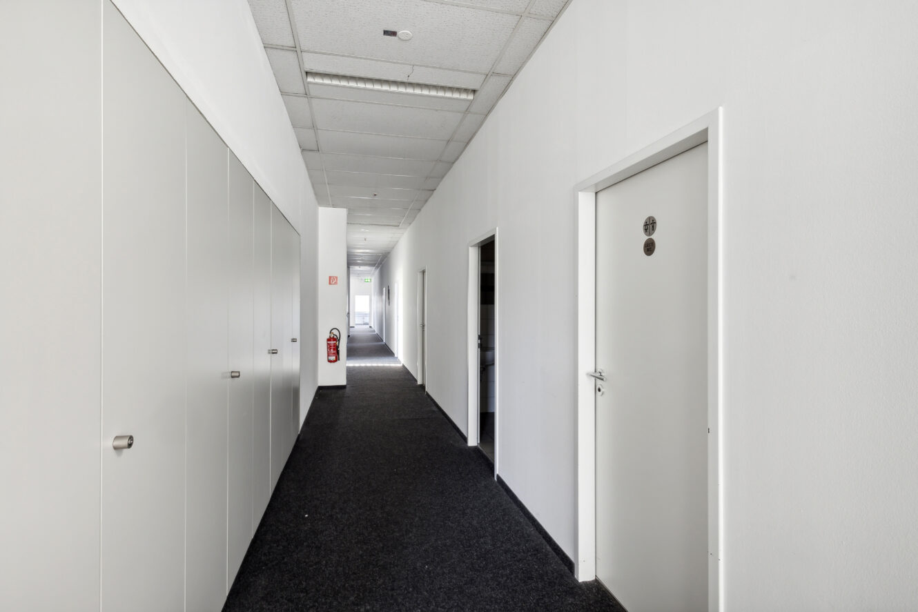 Ein sauberer, leerer Flur in einem Bürogebäude mit weißen Wänden, einem dunklen Teppich, geschlossenen Türen und einem an der Wand angebrachten Feuerlöscher.