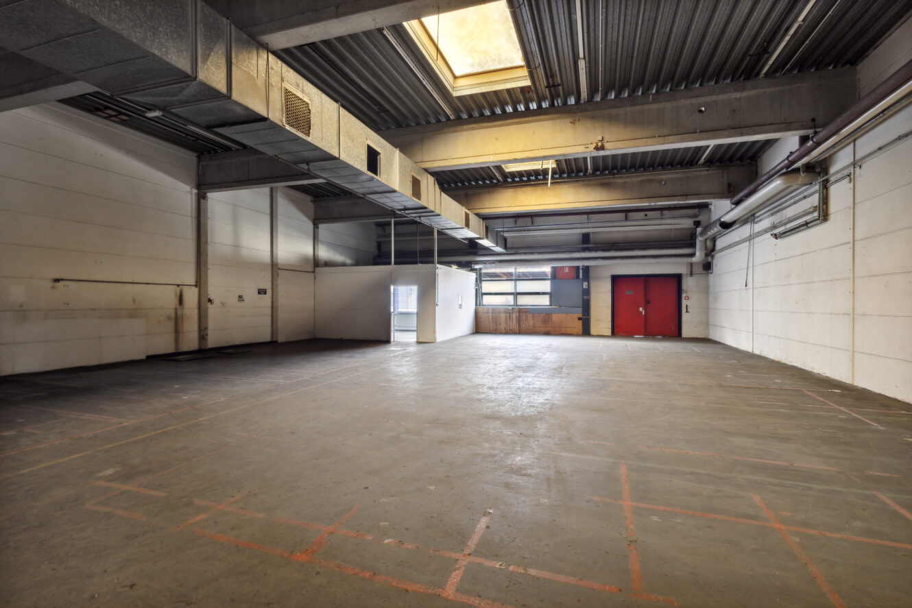 Leeres Industrielager mit Betonboden, weißen Wänden, sichtbaren Deckenbalken, großen roten Flügeltüren und einem Oberlicht, das natürliches Licht hereinlässt.