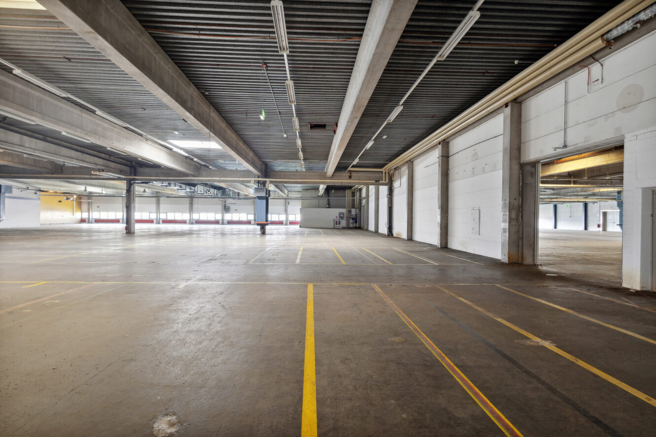 Das Innere eines leeren Industrielagers mit Betonböden, hohen Decken, freiliegenden Balken und markierten gelben Linien auf dem Boden.