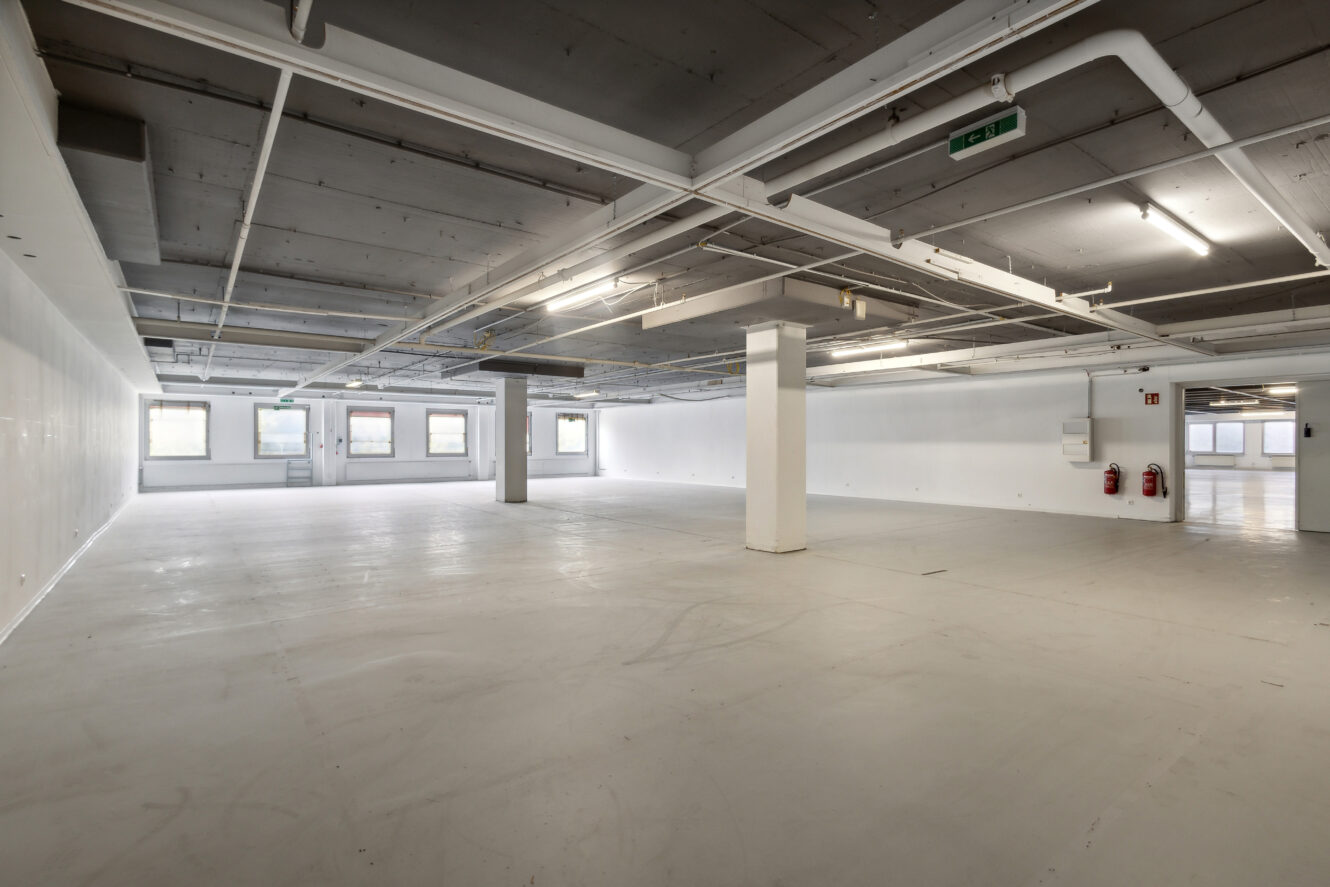Großer leerer Industrieraum mit Betonboden, freiliegenden Deckenrohren, weißen Säulen, Feuerlöschern an der Wand und mehreren Fenstern, die natürliches Licht hereinlassen.