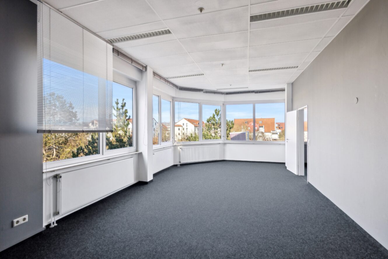 Ein leerer Büroraum mit großen Fenstern, grauem Teppich, weißen Wänden und Blick auf die Bäume und Gebäude draußen.