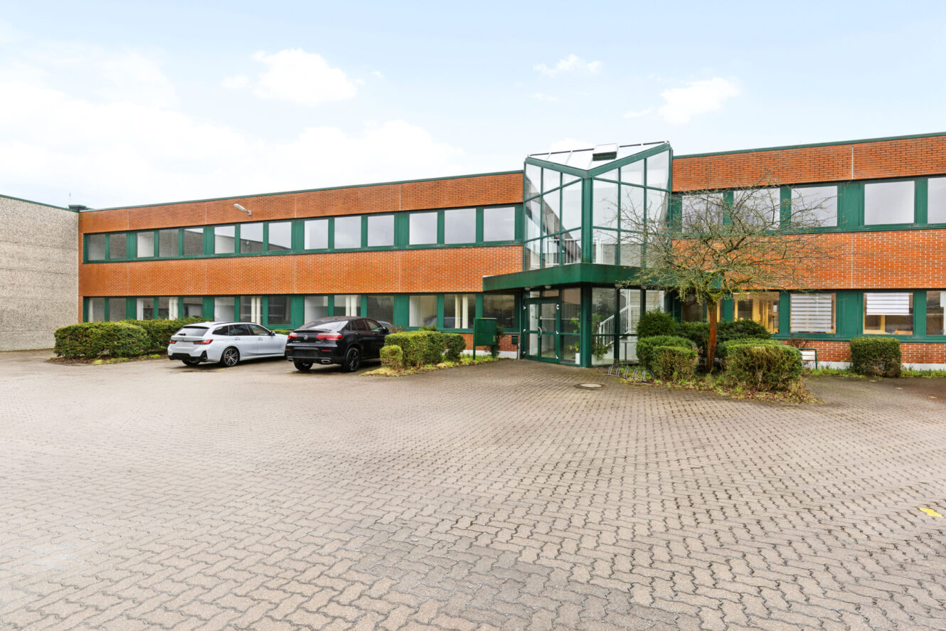 Ein zweistöckiges Bürogebäude mit Backsteinfassade, grün umrahmten Fenstern und einem gläsernen Eingang; davor sind drei Autos auf einem gepflasterten Platz geparkt.