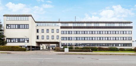 Ein vierstöckiges, rechteckiges Bürogebäude mit großen Fenstern, weißer Fassade und einer Einfahrt, die unter einem Abschnitt hindurchführt, an einem klaren Tag von der anderen Straßenseite aus gesehen.