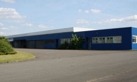 Ein großes, blaues Industrielager mit mehreren Fenstern und Türen, umgeben von einer gepflasterten Fläche und etwas Grün unter einem teilweise bewölkten Himmel.