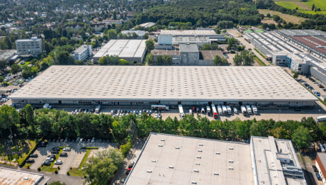 Luftaufnahme eines großen Lagerhauses mit mehreren Laderampen, umgeben von geparkten Lastwagen, Autos und Industriegebäuden, mit einem Waldgebiet im Hintergrund.