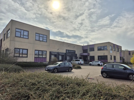 Ein modernes zweistöckiges Bürogebäude mit mehreren geparkten Autos davor, unter einem bewölkten Himmel mit sichtbarer Sonne. Büsche säumen den Vordergrund.