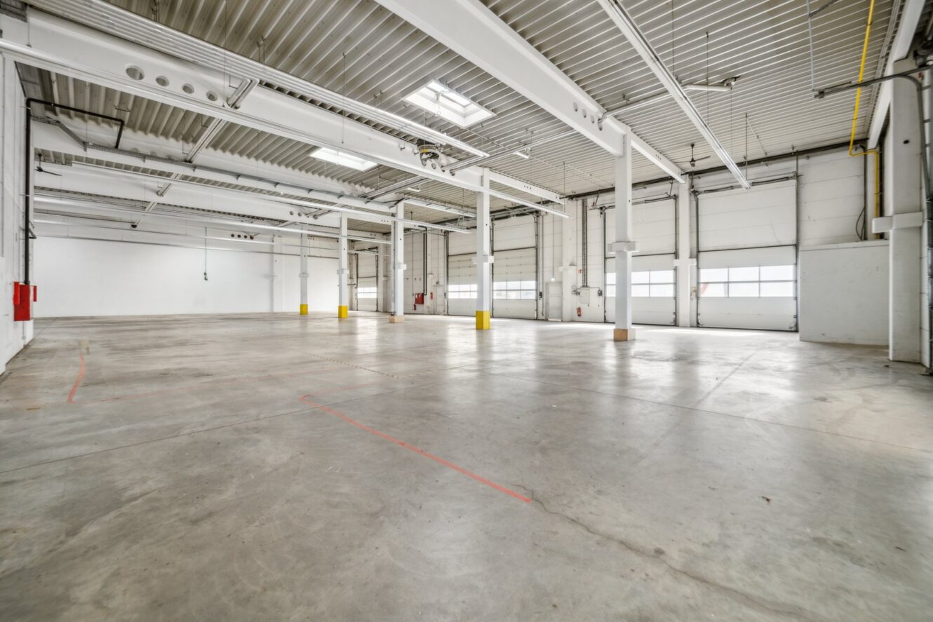 Leeres Industrielager mit Betonboden, hoher Decke, großen Fenstern, Oberlichtern, freiliegenden Rohren und Stützpfeilern.