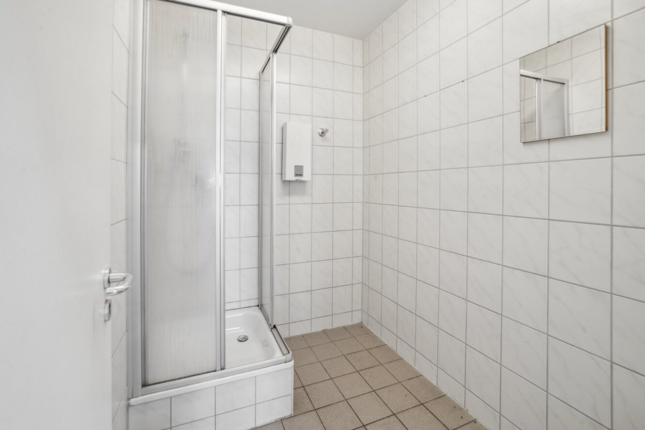 Ein weiß gefliestes Badezimmer mit einer Eckdusche, einem Seifenspender an der Wand, einem kleinen Spiegel und einem gefliesten Boden.