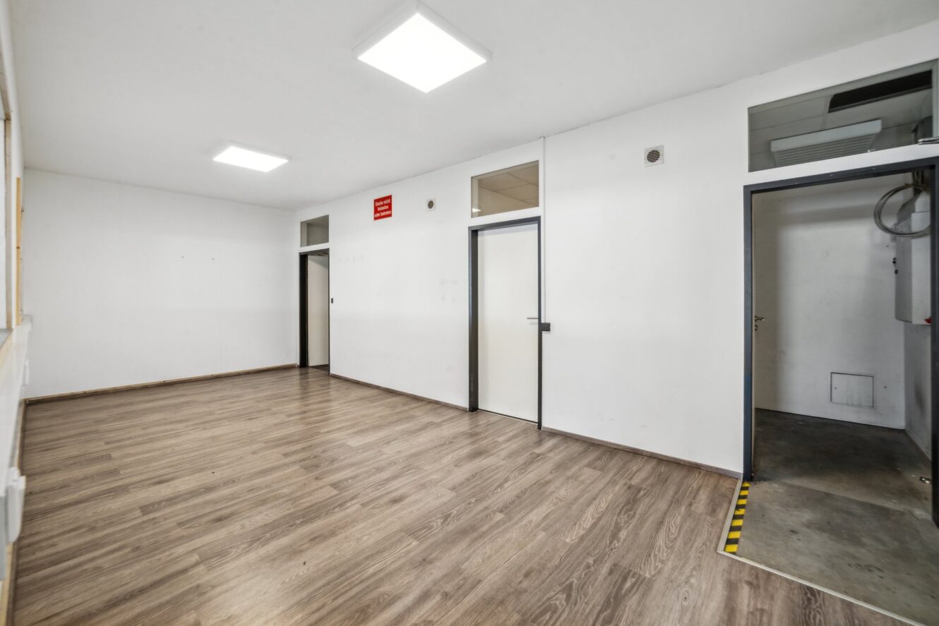 Leerer Raum mit weißen Wänden, Holzfußboden, drei Türen, Deckenbeleuchtung und einem kleinen roten Schild über einer Tür.