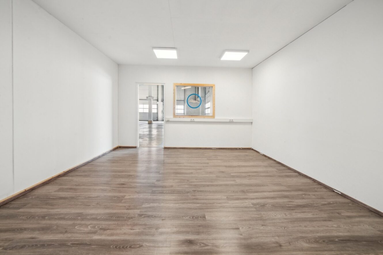 Leerer Raum mit weißen Wänden, Holzfußboden, einem großen Innenfenster, Leuchtstoffröhren an der Decke und einer Tür, die zu einem anderen Raum führt.