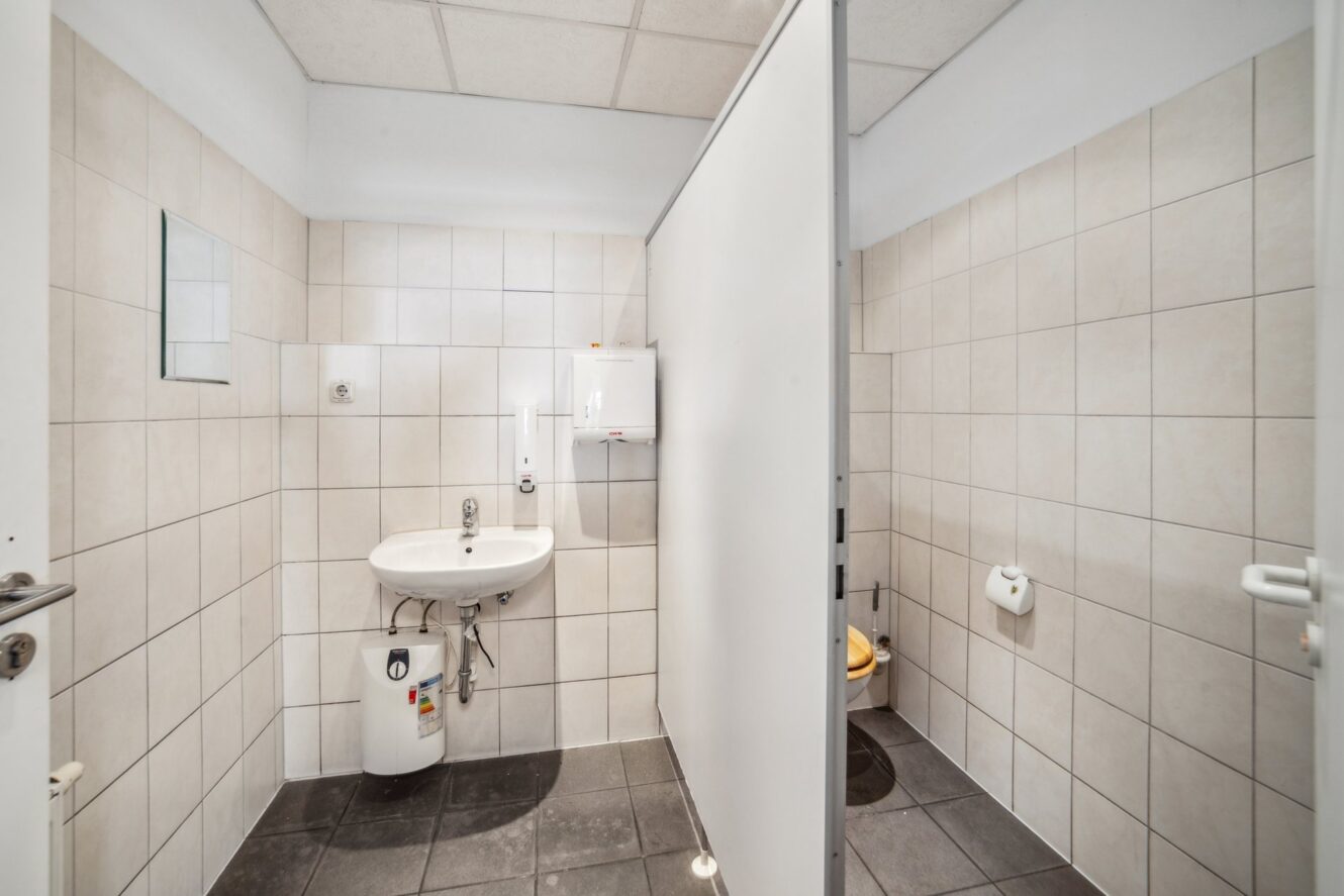 Eine öffentliche Toilette mit gefliesten Wänden, einem Waschbecken mit Spiegel, Seifenspender und Papierhandtuchhalter auf der linken Seite und einer Toilette in einer separaten Kabine auf der rechten Seite.