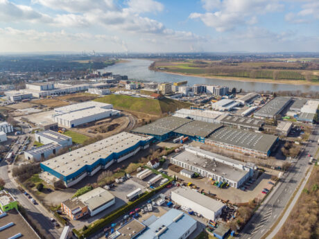 Luftaufnahme eines Industriegebiets mit Lagerhäusern, Bürogebäuden, Straßen und einem Fluss im Hintergrund bei teilweise bewölktem Himmel.