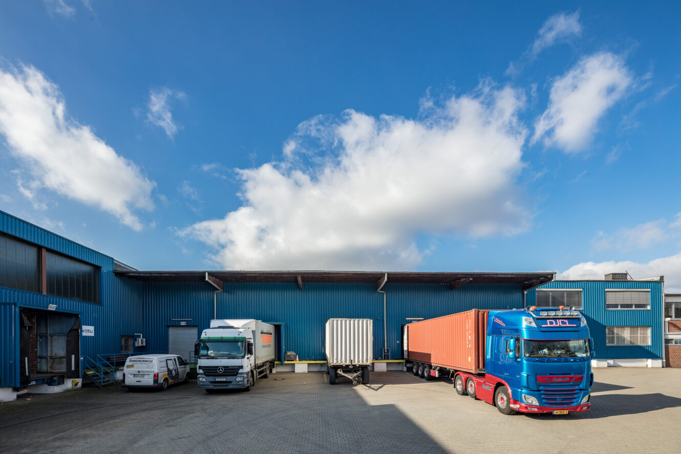 Eine Laderampe mit blauen Industriegebäuden, drei Lastwagen - ein roter und zwei weiße -, die nebeneinander geparkt sind, und ein teilweise bewölkter Himmel über dem Kopf.