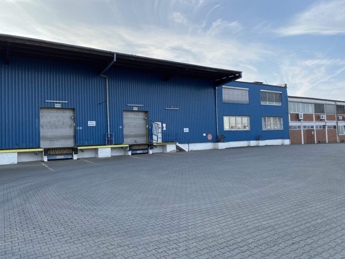 Ein blaues industrielles Lagergebäude mit zwei geschlossenen Laderampen, einem leeren gepflasterten Parkplatz und einem teilweise bewölkten Himmel über uns.