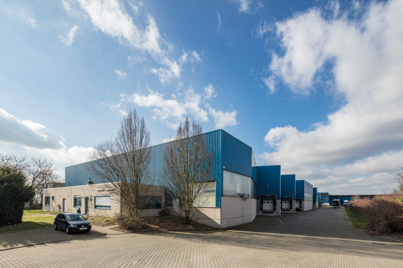 Großes Industrielager mit blauen Metallverkleidungen und Laderampen, angrenzend an einen Parkplatz mit einem geparkten Auto, unter einem teilweise bewölkten Himmel.