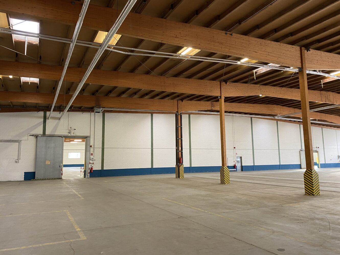 Geräumiges, leeres Lagerhaus mit Betonboden, Holzbalken, weißen Ziegelwänden und gelb gestreiften Säulenschutzvorrichtungen.