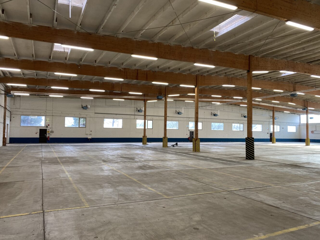 Große leere Lagerhalle mit Betonboden, Holzbalken, Leuchtstoffröhren und Fenstern an den Wänden. Gelbe Sicherheitsbarrieren umschließen einige Stützpfeiler.