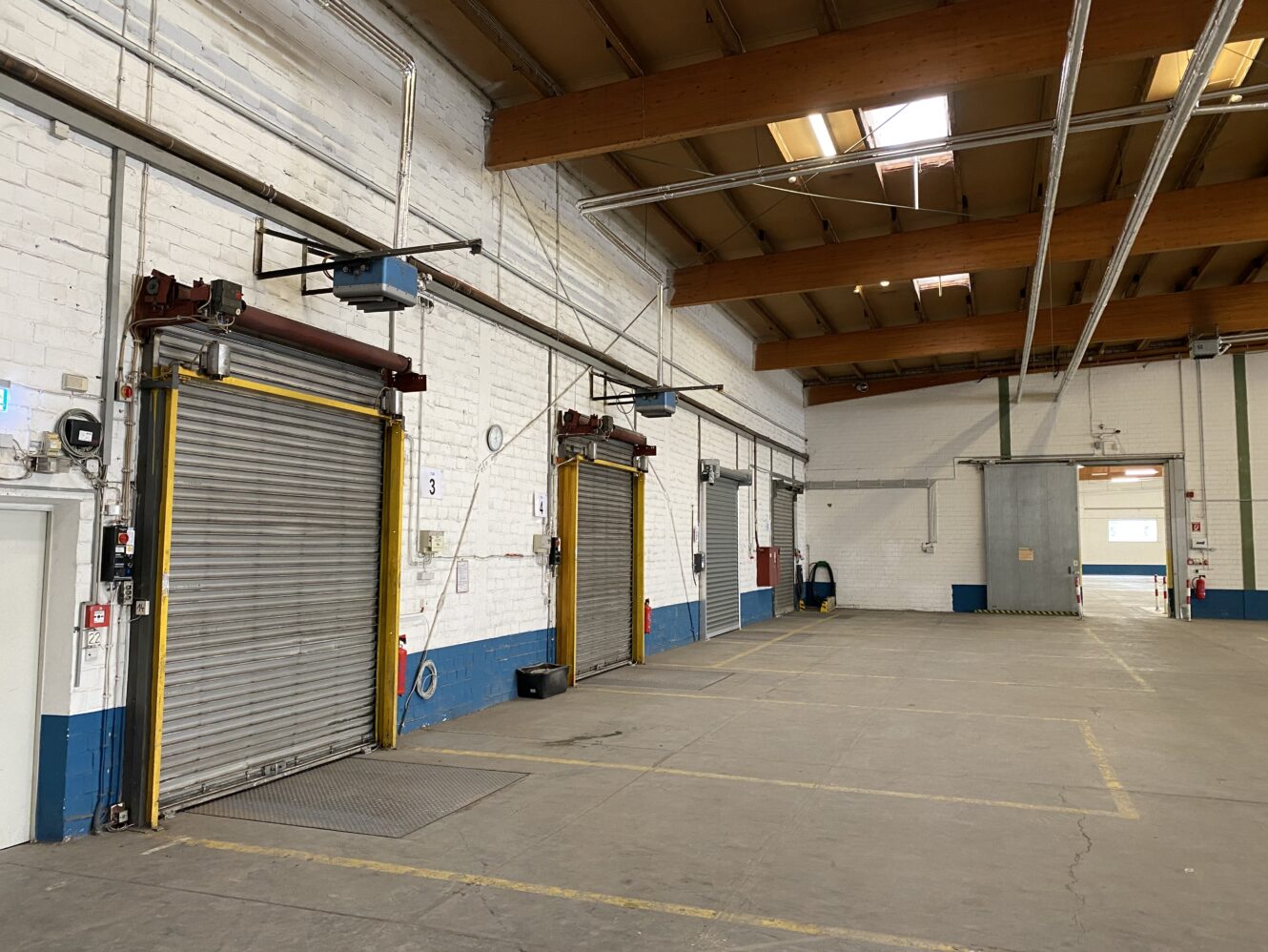 Leeres Lagerhaus mit zwei geschlossenen Laderaumtoren, freiliegenden Balken, weißen Ziegelwänden und Betonboden. Gelbe und blaue Sicherheitsmarkierungen sind an den Wänden und Türen zu sehen.