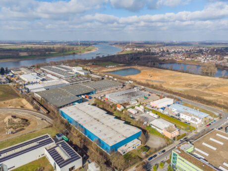 Luftaufnahme eines Industriegebiets mit Lagerhallen und Fabriken in der Nähe eines Flusses, mit einigen Freiflächen und einem Wohngebiet im Hintergrund unter einem teilweise bewölkten Himmel.